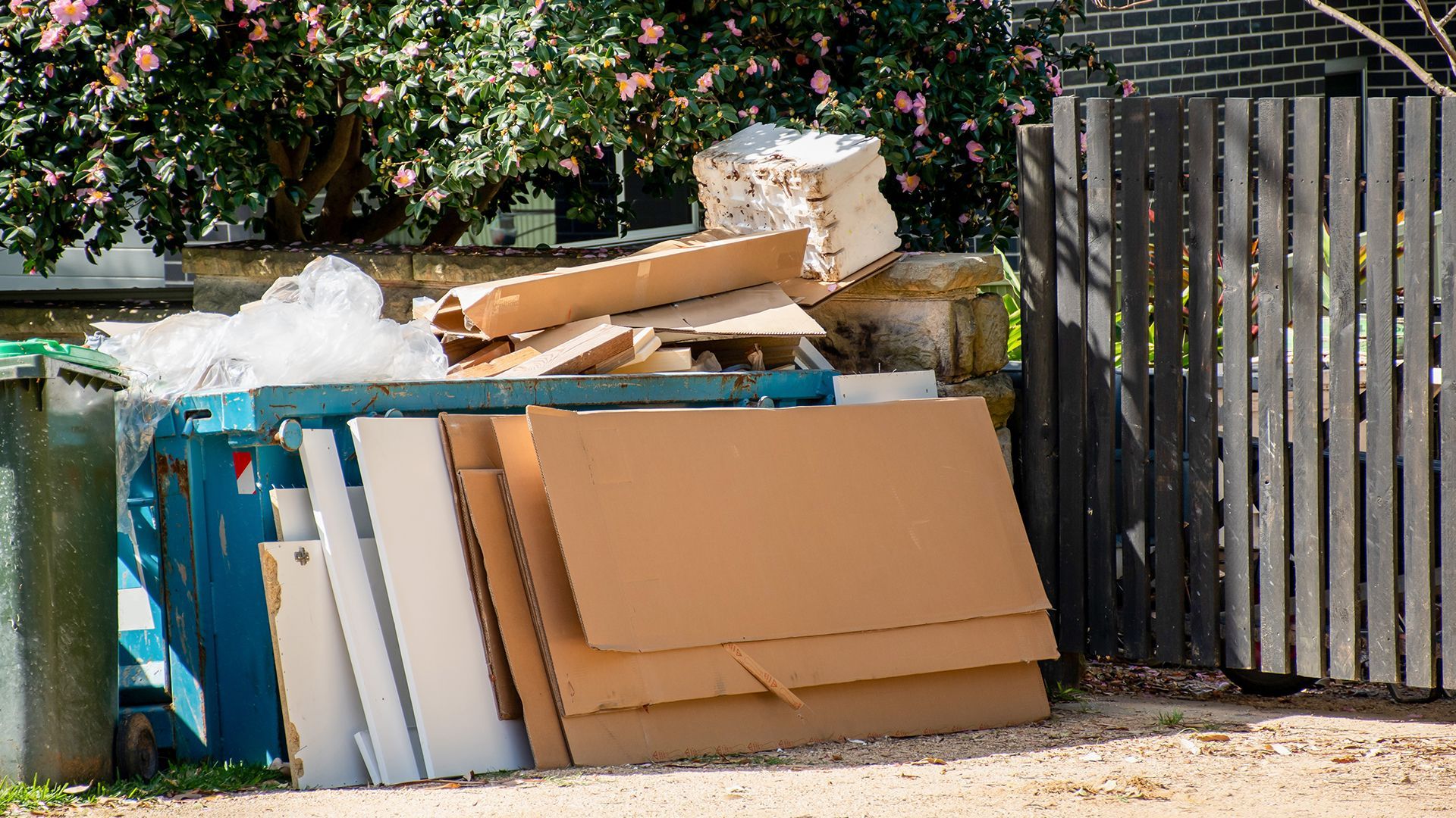 Overflowing blue recycling bin with stacked cardboard boxes next to a wooden fence.