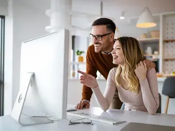 Couple smiling, looking at a computer screen. Man points, woman's arm around her. Kitchen setting.