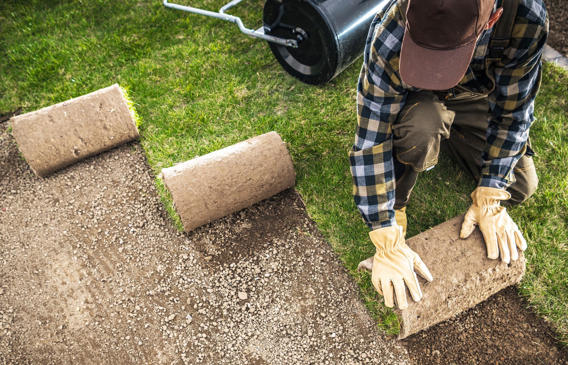 Person laying tan carpet tiles on a lawn with a roller nearby