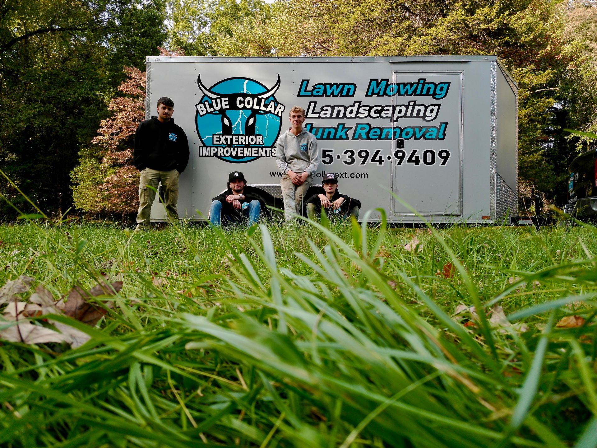 People pose in front of a white trailer for a lawn care business, in a grassy area with trees.