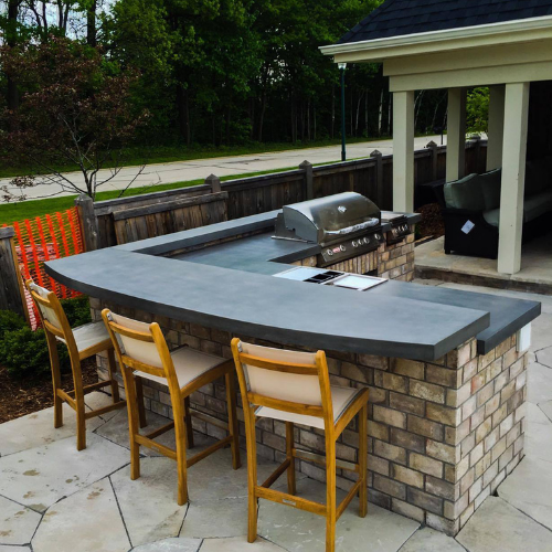 A brick outdoor kitchen with a concrete countertop, a grill and stools.
