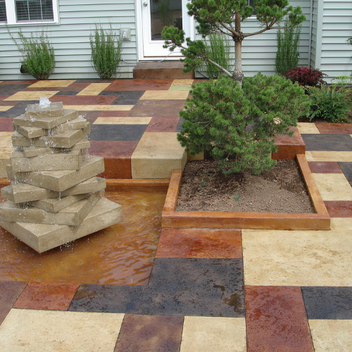 A stained concrete patio with a fountain and a tree in front of a house