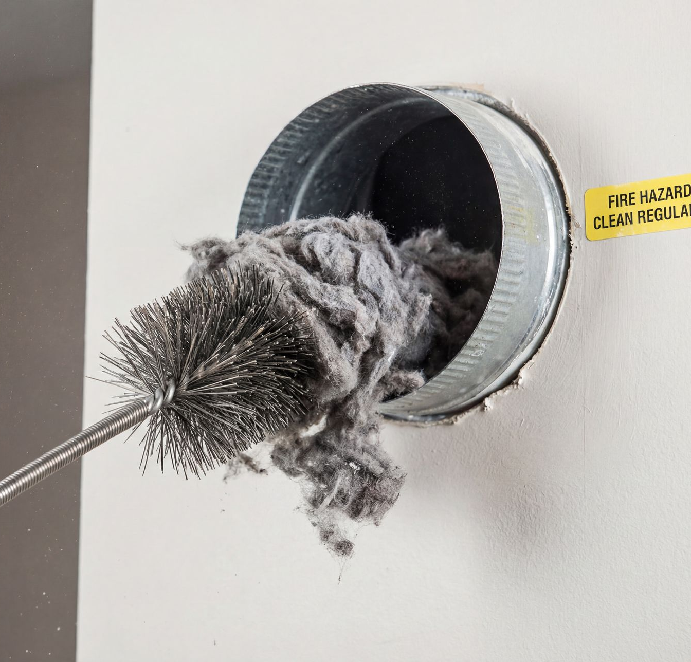 Person cleaning lint from a wall vent with a brush and vacuum hose