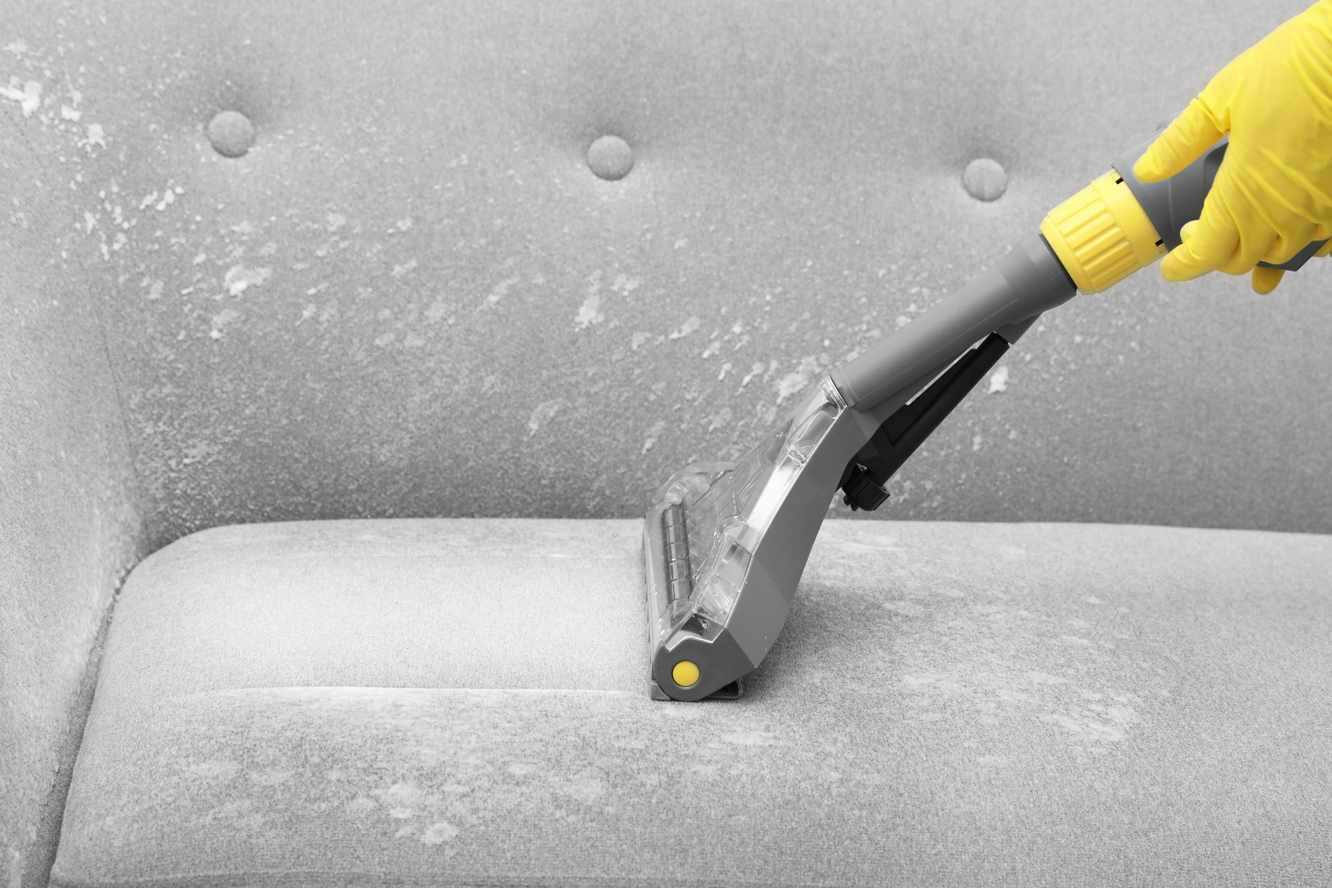 Vacuum nozzle cleaning a gray couch, with a yellow-gloved hand visible.
