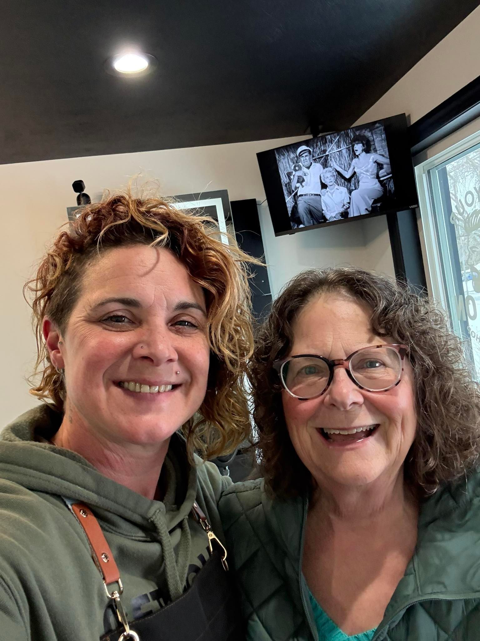 Two smiling women indoors; one in green hoodie, the other with glasses, arm around the first. Black and white photo on TV.
