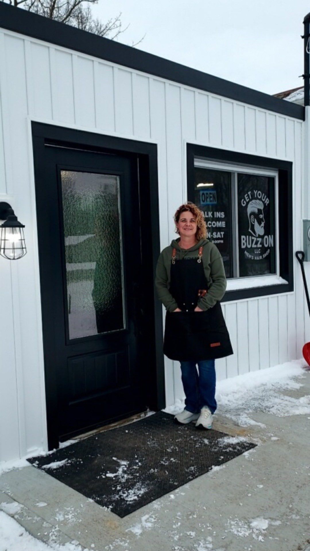 Woman in black apron stands in front of a white building with black door and window; snow on the ground.
