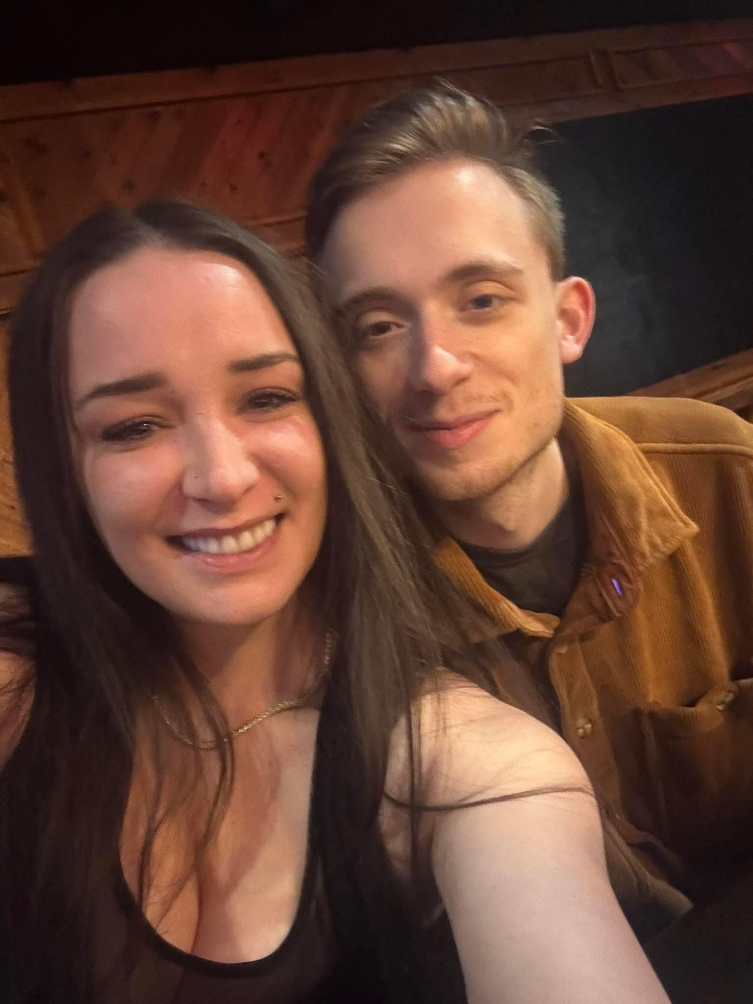 Woman and man smile close together. Woman has long brown hair, man wears a tan jacket. Dim lighting, indoor setting.