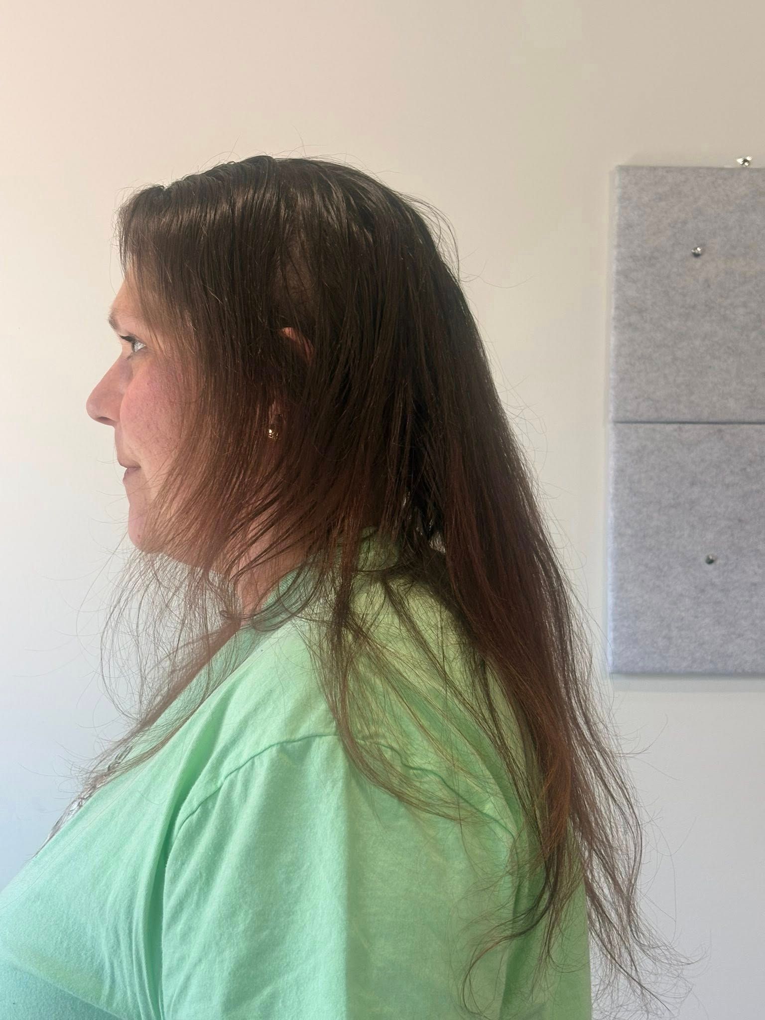 Woman in a green shirt with long, brown hair, looking left. Standing in front of a white wall.