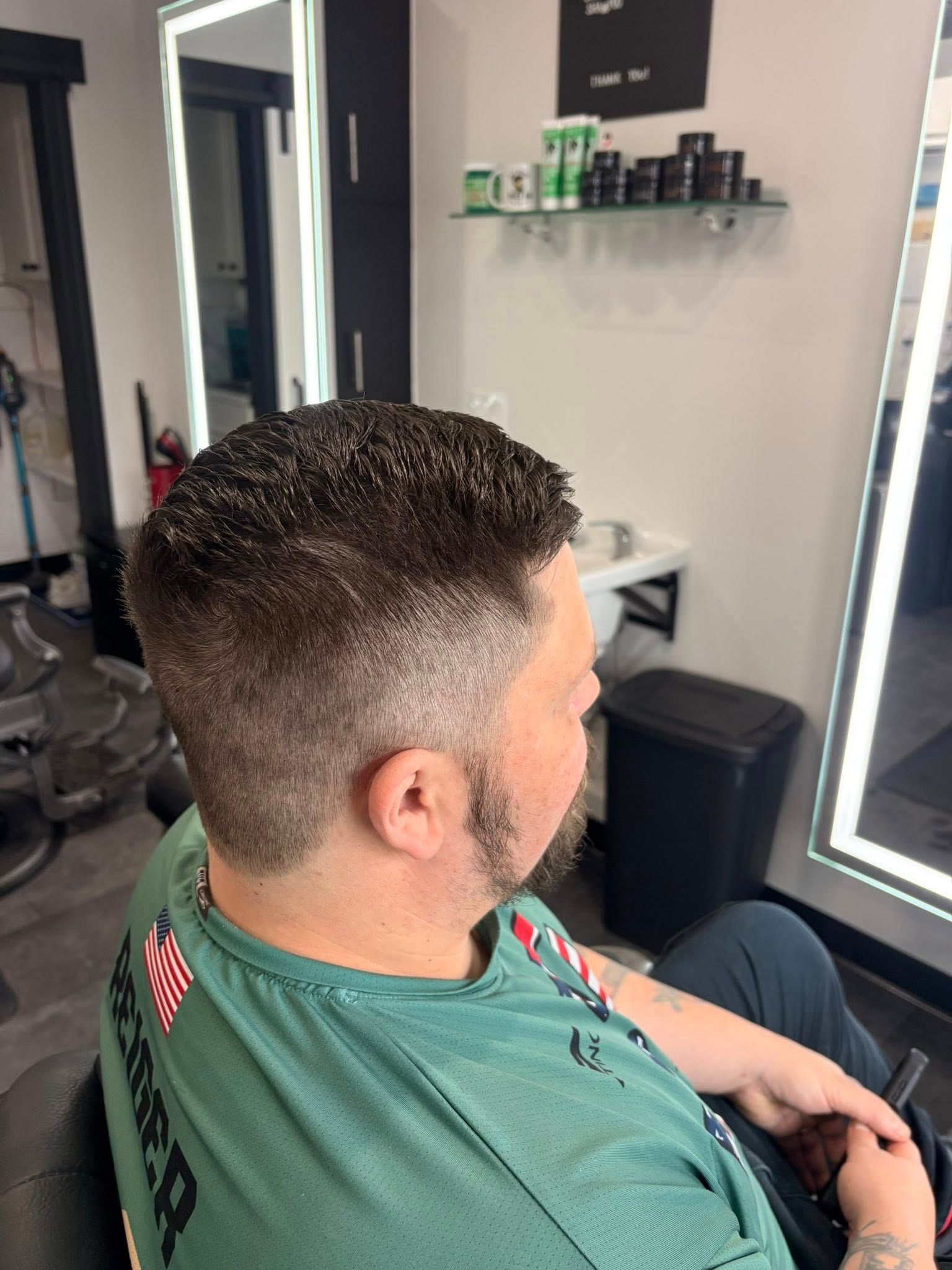 Man getting a haircut at a salon, with dark hair on top and faded sides. He wears a green shirt.