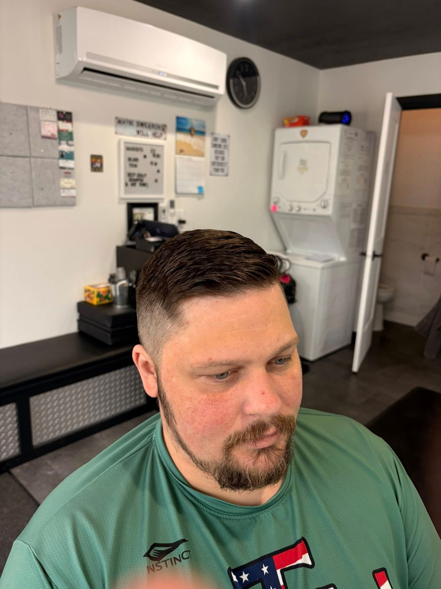 Man with a fresh haircut in a barbershop; black side table, white appliances, and air conditioning unit in the background.