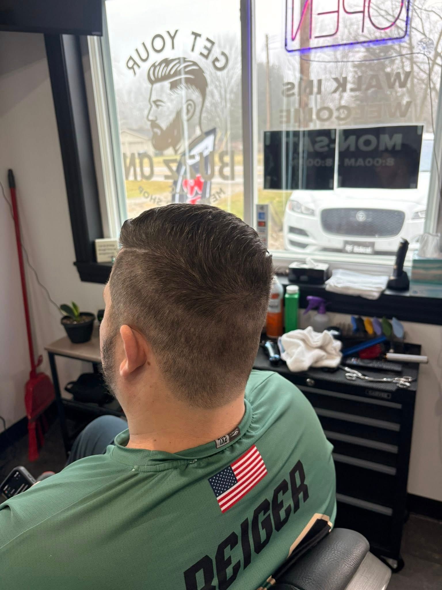 Person in barber chair getting a haircut.  Barber shop interior.  He is wearing a green shirt with an American flag.