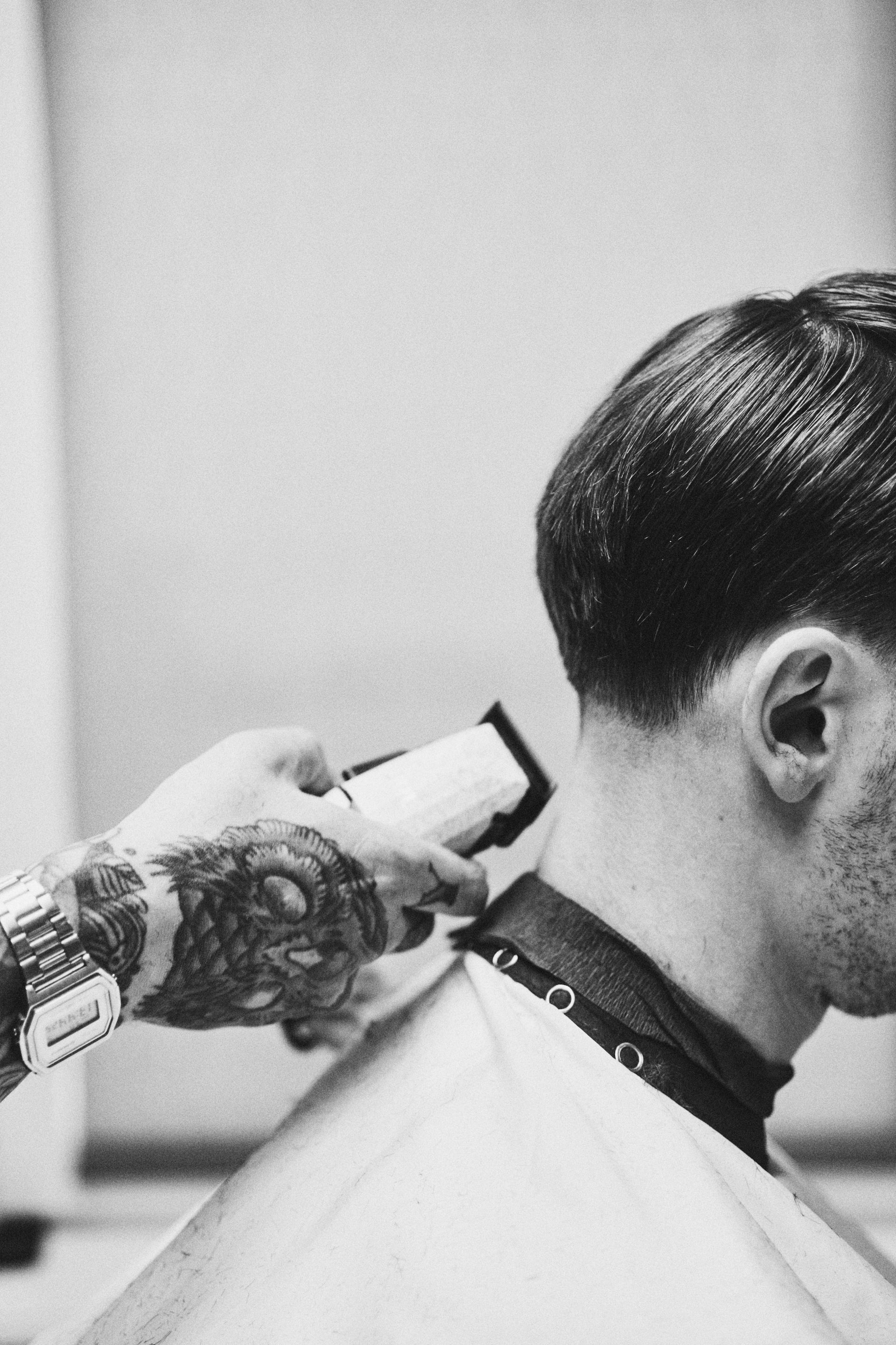 Barber giving a haircut using clippers; close-up of neck and tattooed arm.