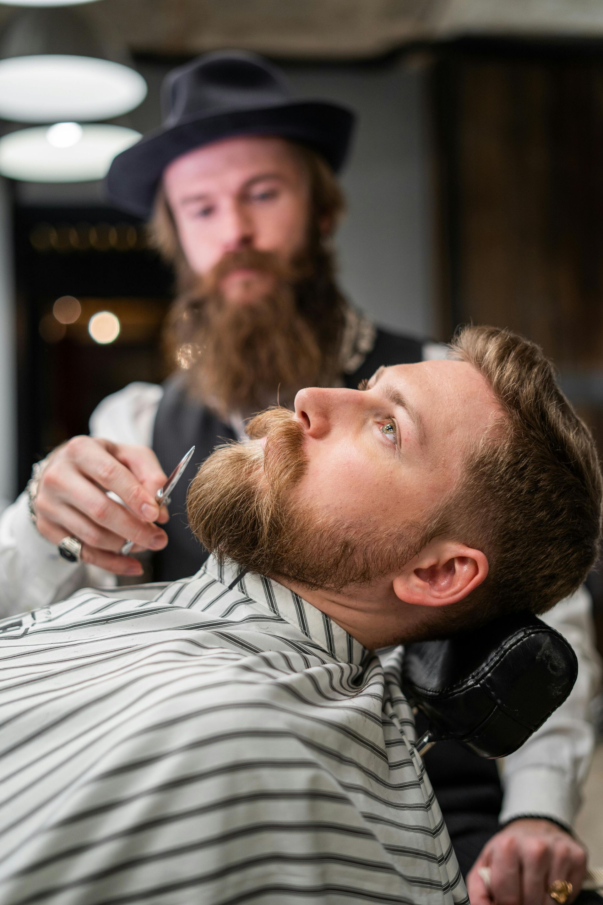 Barber trimming a customer's beard in a barbershop. The customer is reclining, the barber is standing with a black hat on.