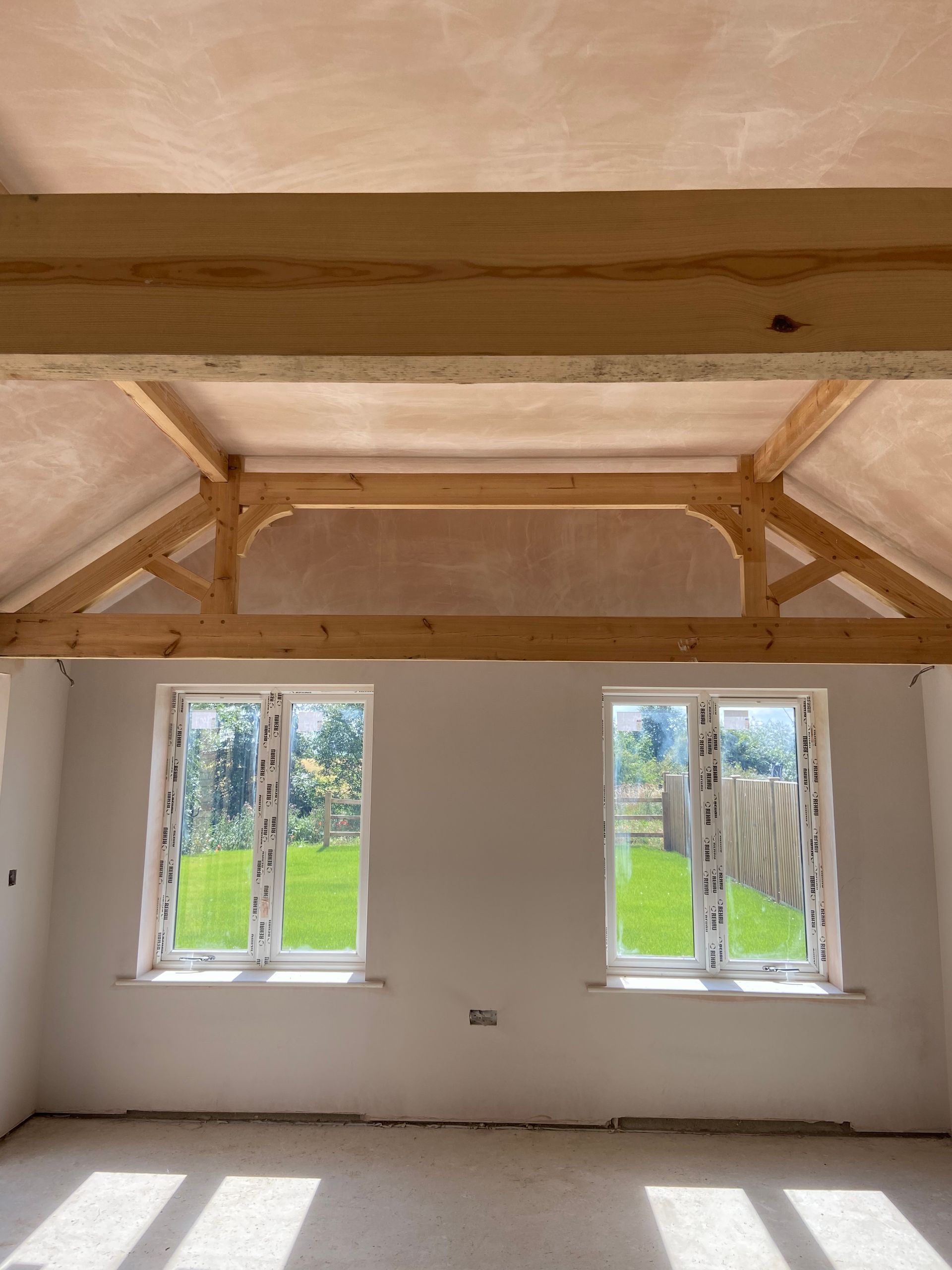 Interior view of a room with exposed wooden beams, two windows overlooking a green lawn, and plaster walls.
