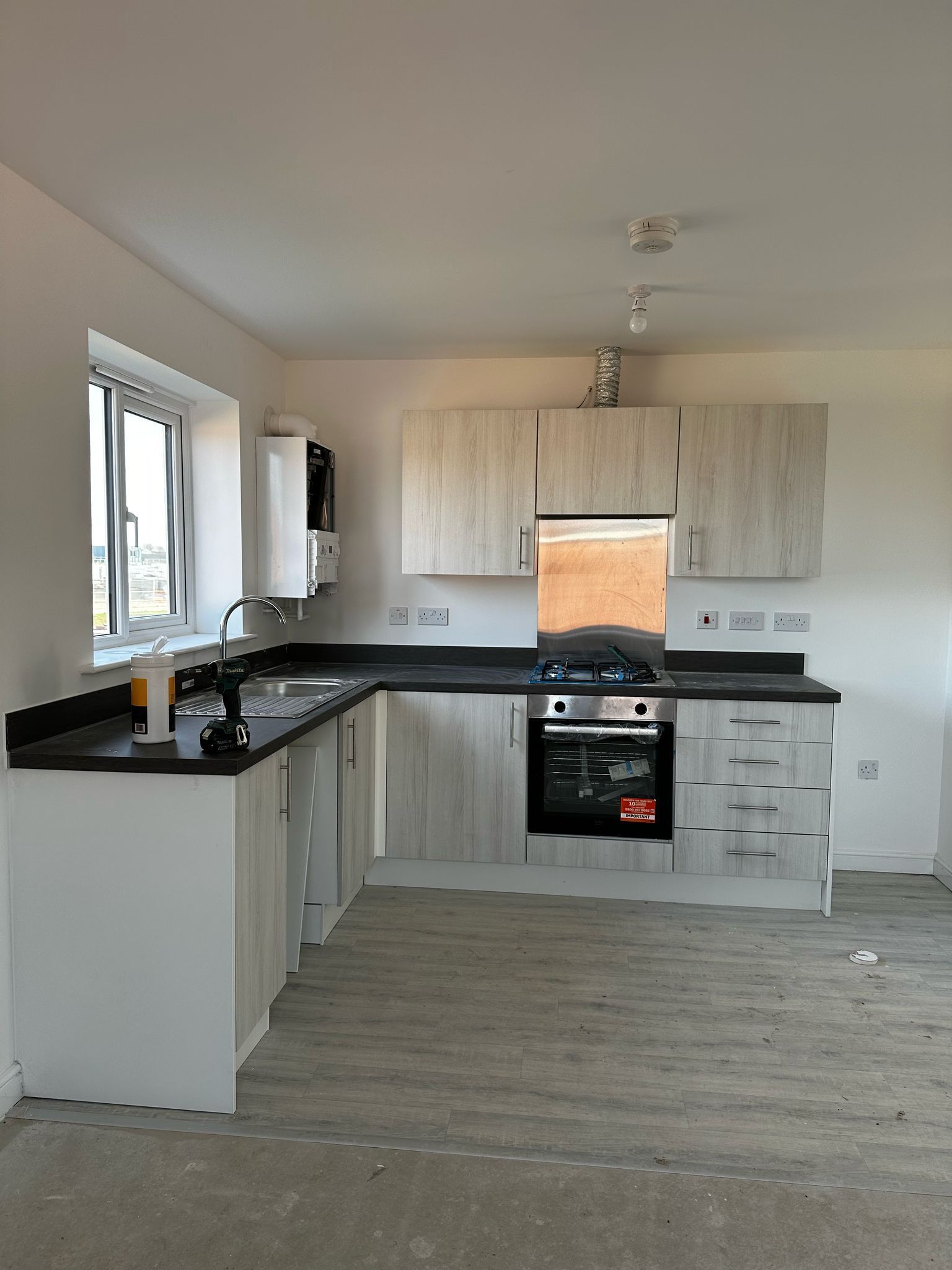 L-shaped kitchen with light wood cabinets, black countertops, and stainless steel appliances in a room with bare flooring.