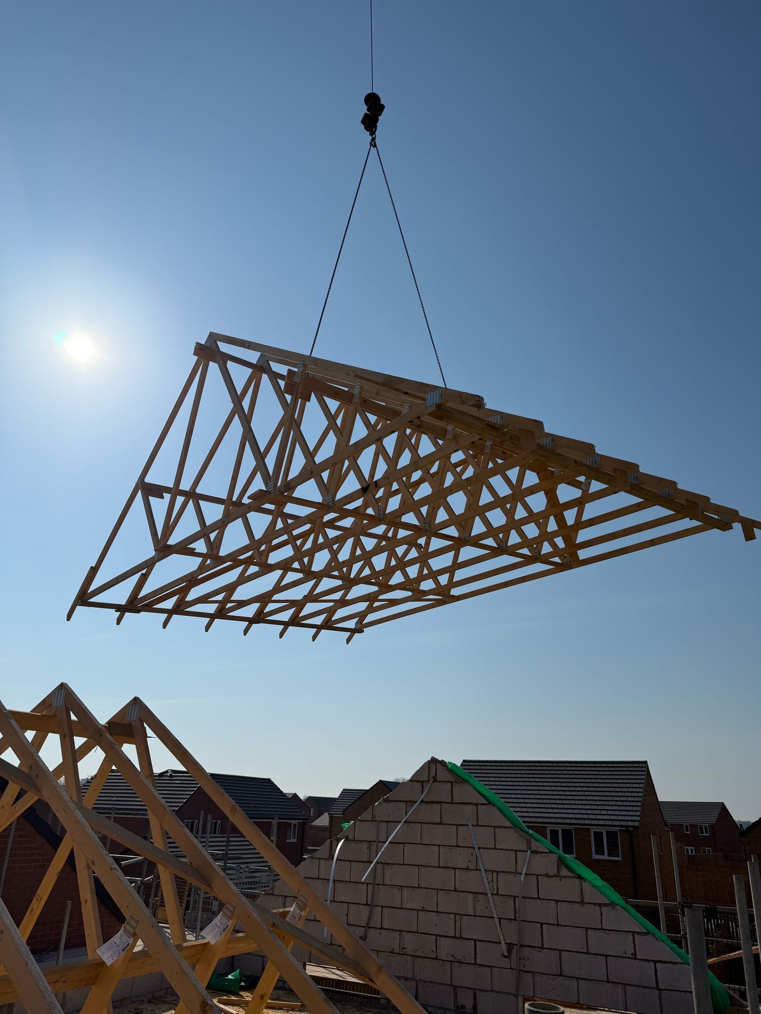 Crane lifting a wooden roof truss into place on a building site under a bright blue sky.