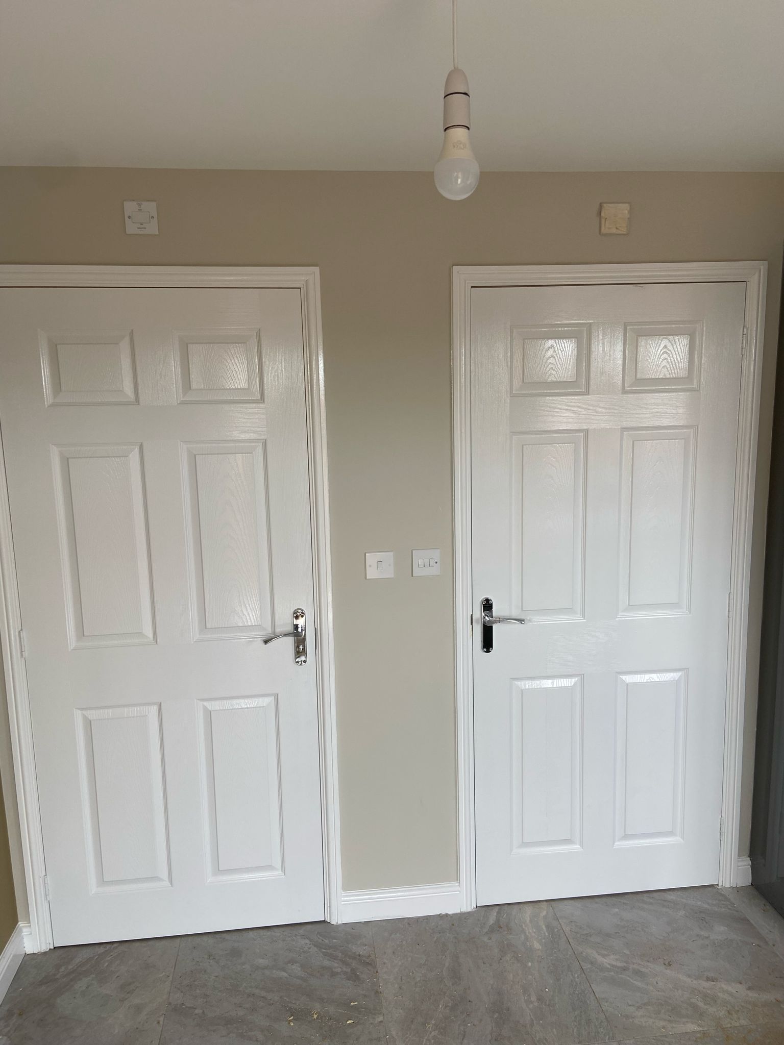 Two white doors, wall with switches and a light bulb hanging from the ceiling.