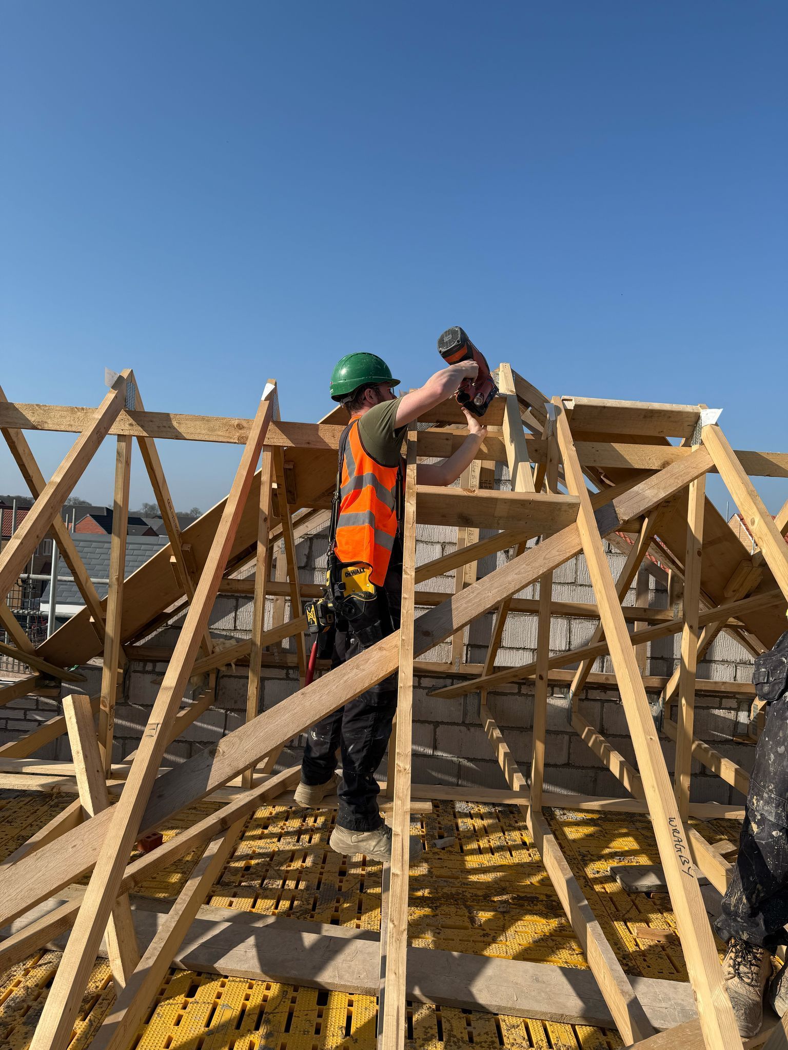 Construction worker on a roof, using a power drill. He is wearing a hard hat and safety vest.