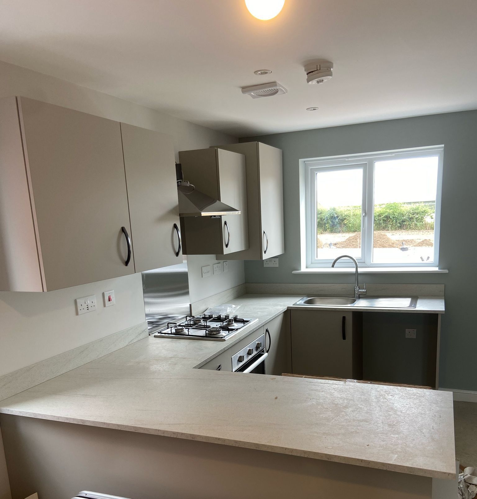 U-shaped kitchen with beige cabinets, countertops, and light blue wall. Stainless steel appliances and sink by a window.