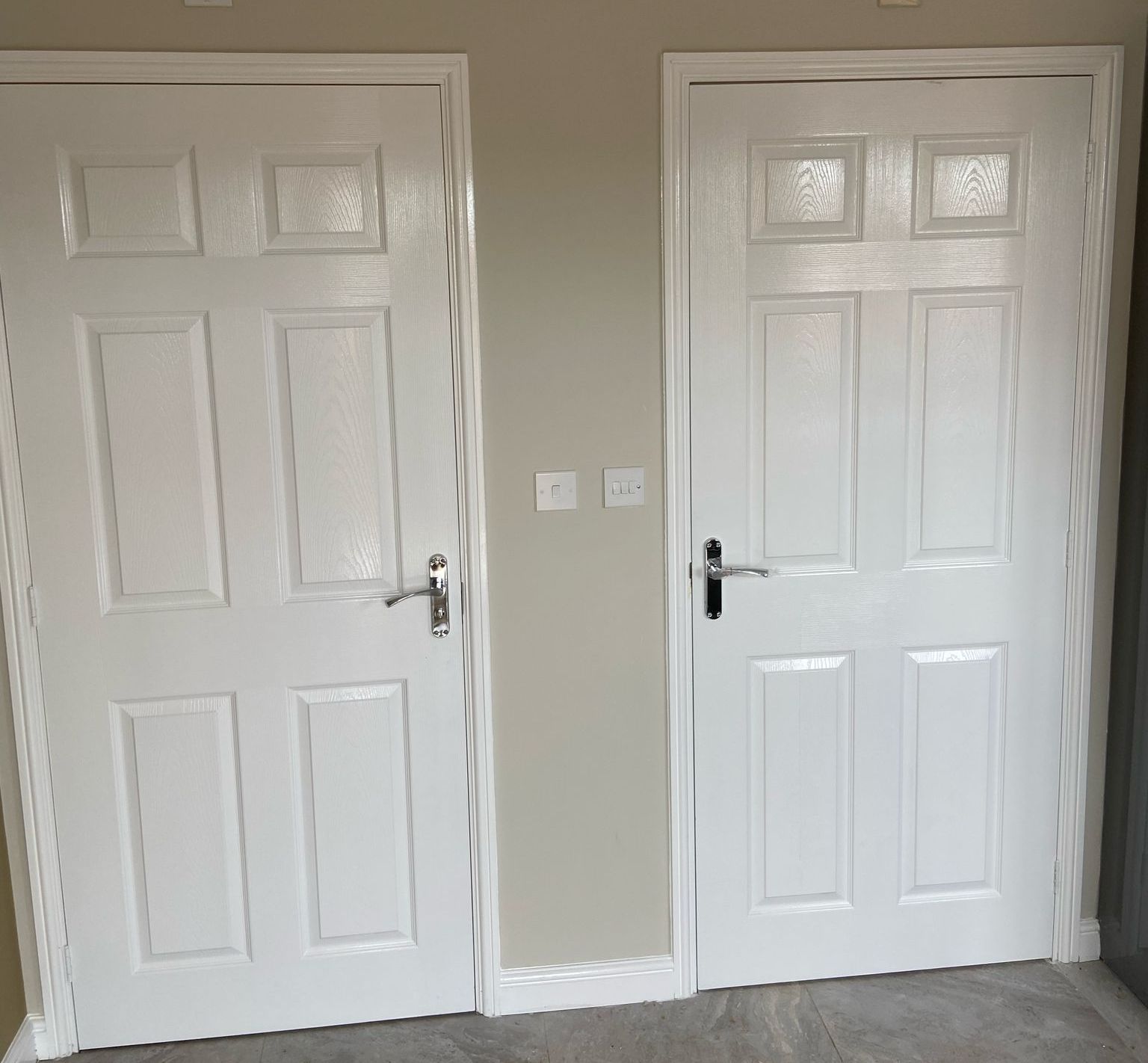 Two white six-panel doors side-by-side on a light gray wall. One has a silver handle, the other black.