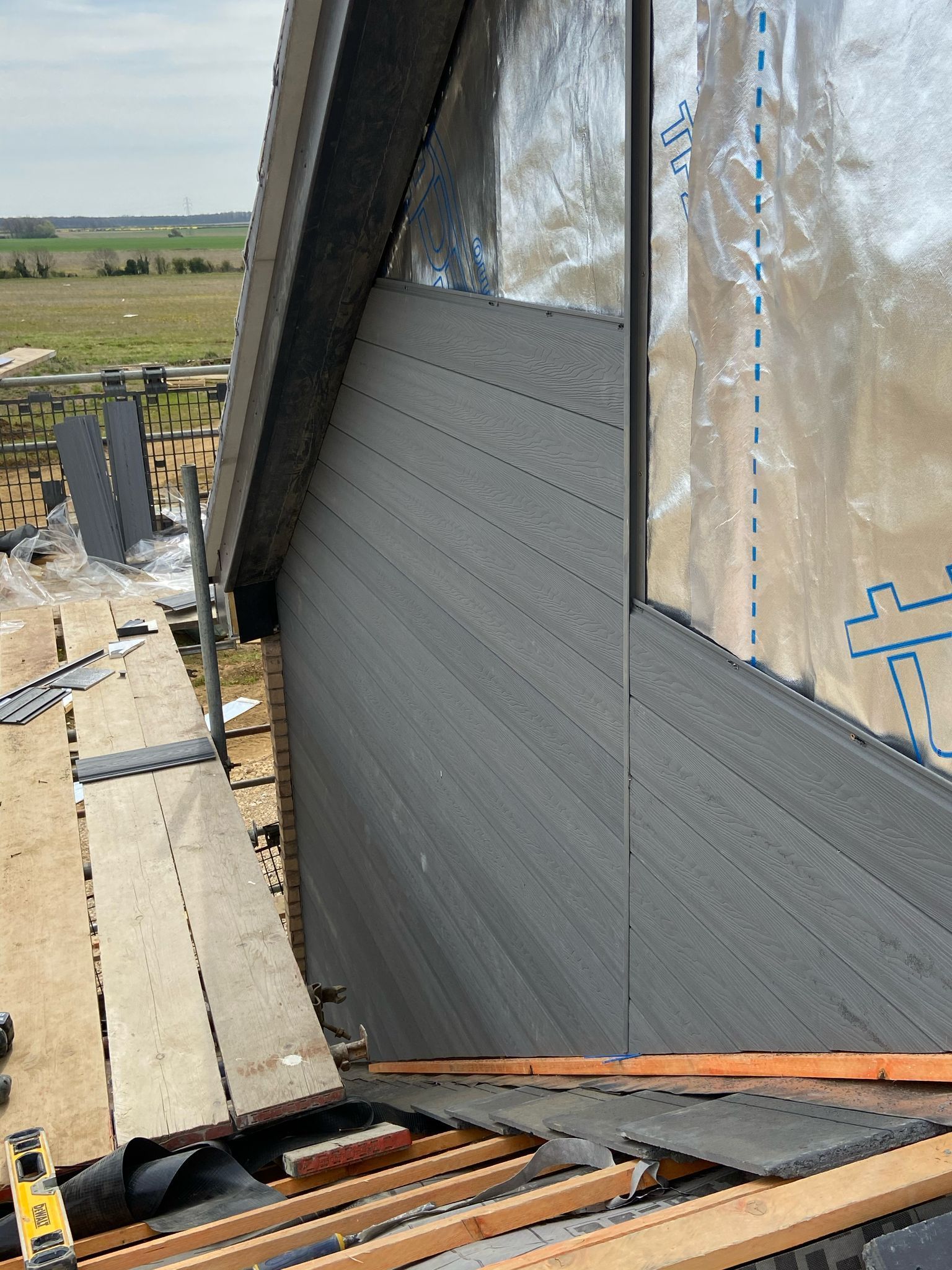 Construction of a building's exterior. Grey siding is installed on angled wall. Wood scaffolding and foil sheathing visible.