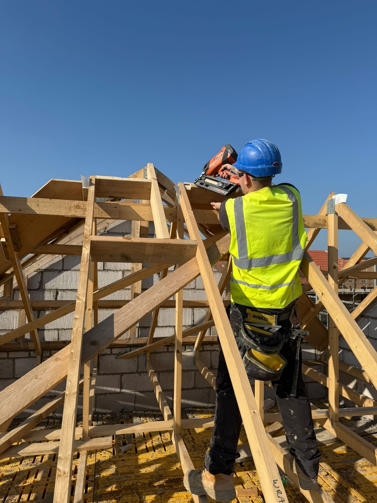 Construction worker on roof using a power saw, wearing safety gear, under a blue sky.