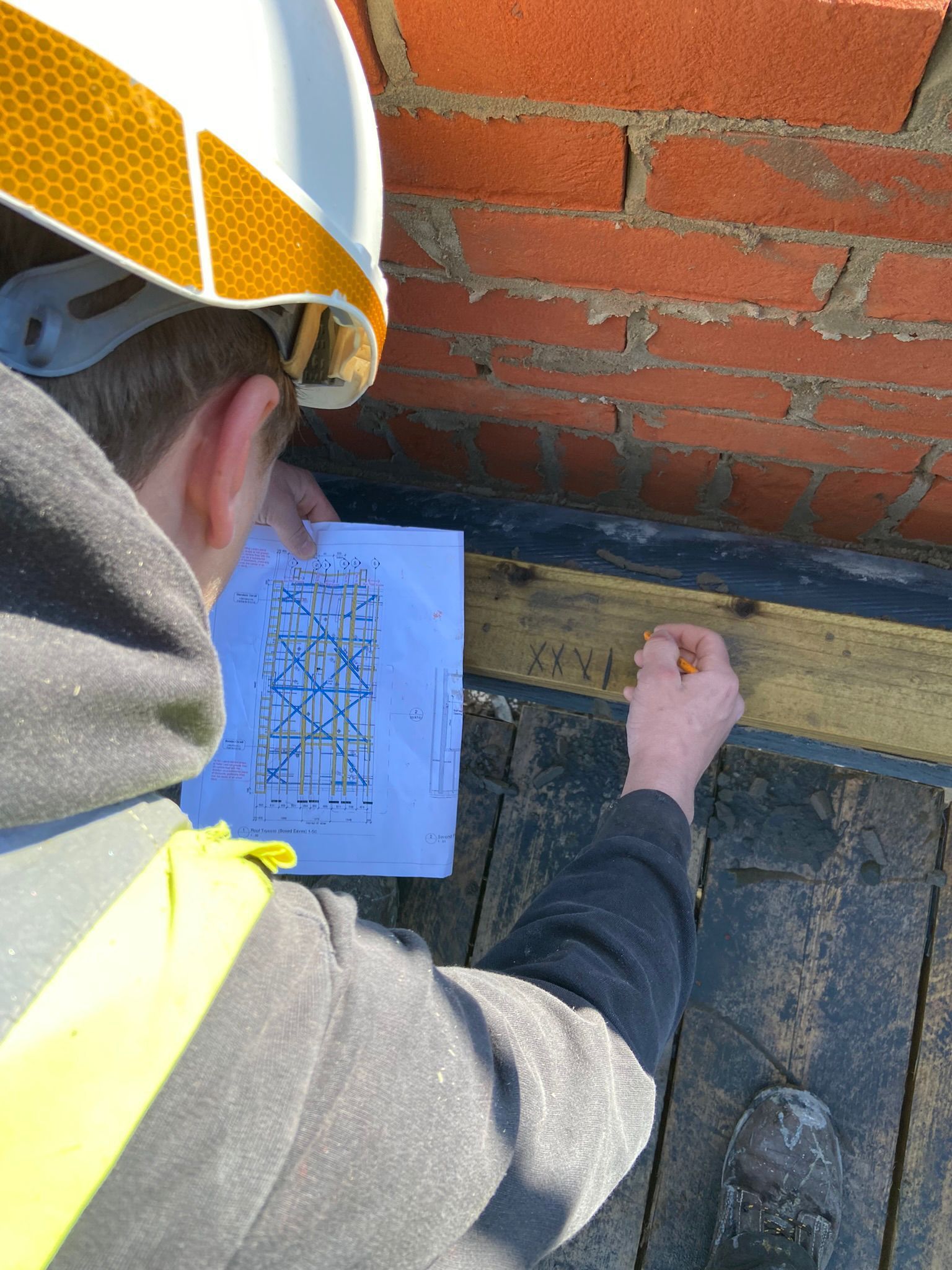 Person in hard hat examining blueprint on a roof near brick wall, marking with pencil.