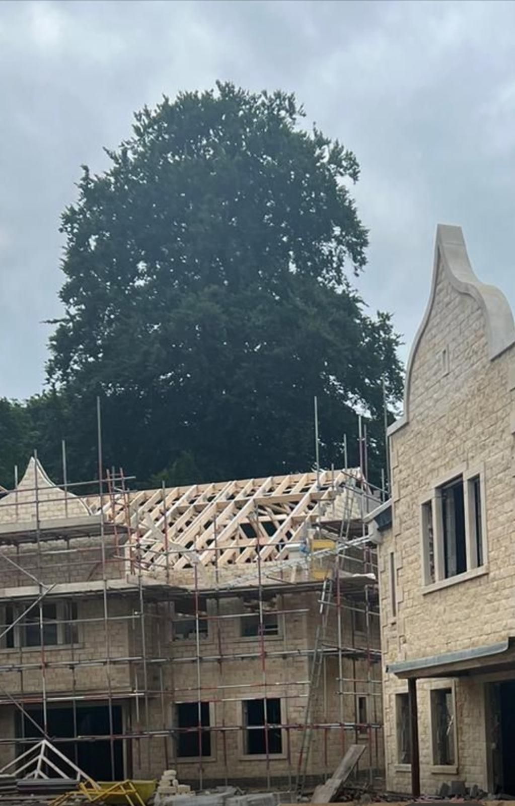 Building under construction with scaffolding, incomplete roof, light-colored stone, and a large tree in the background.