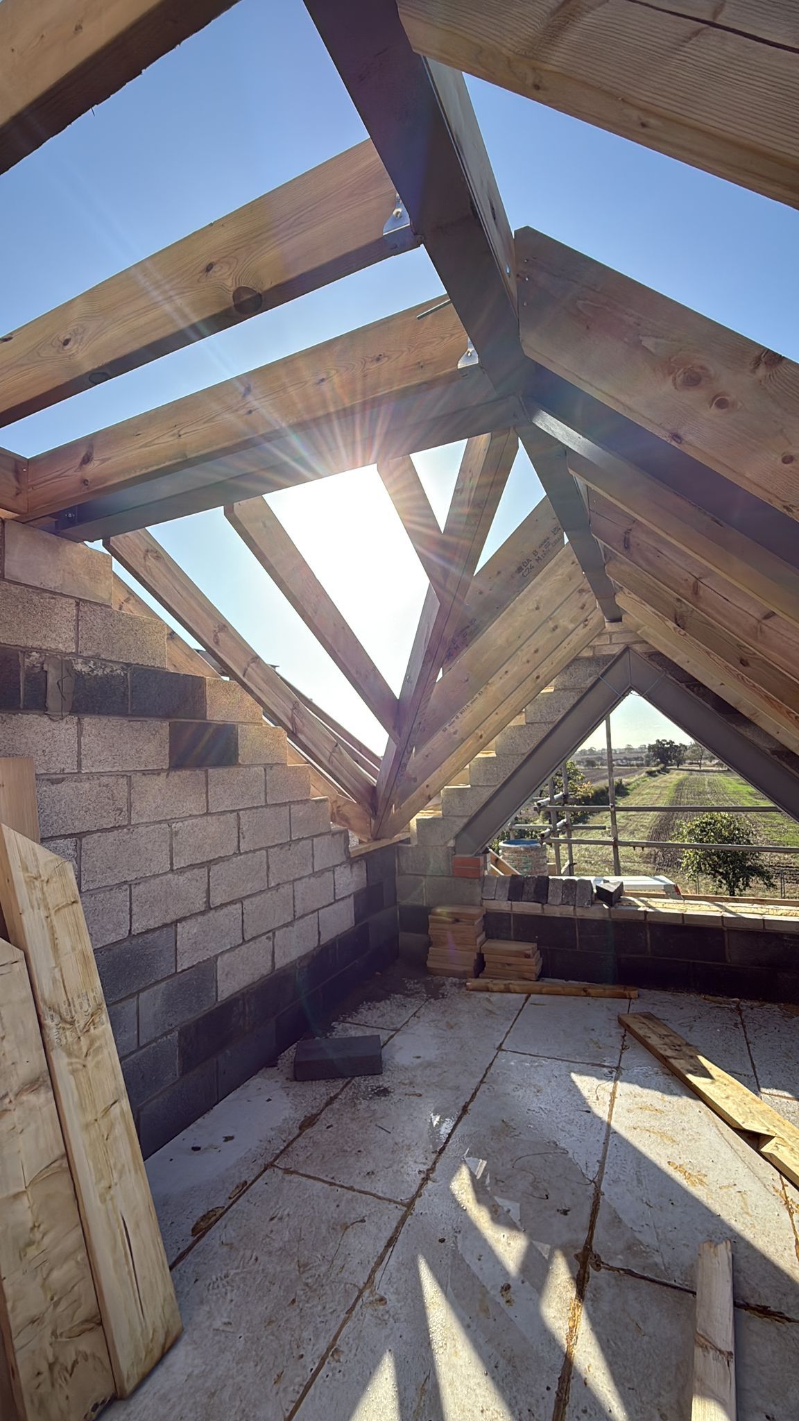 Interior view of a building under construction, featuring wooden rafters and brick walls, with the sun shining through.