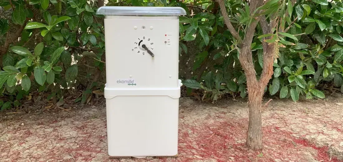 Scatola elettrica color crema con fori di proiettile sulla parte anteriore, albero di fronte e fogliame verde sullo sfondo.