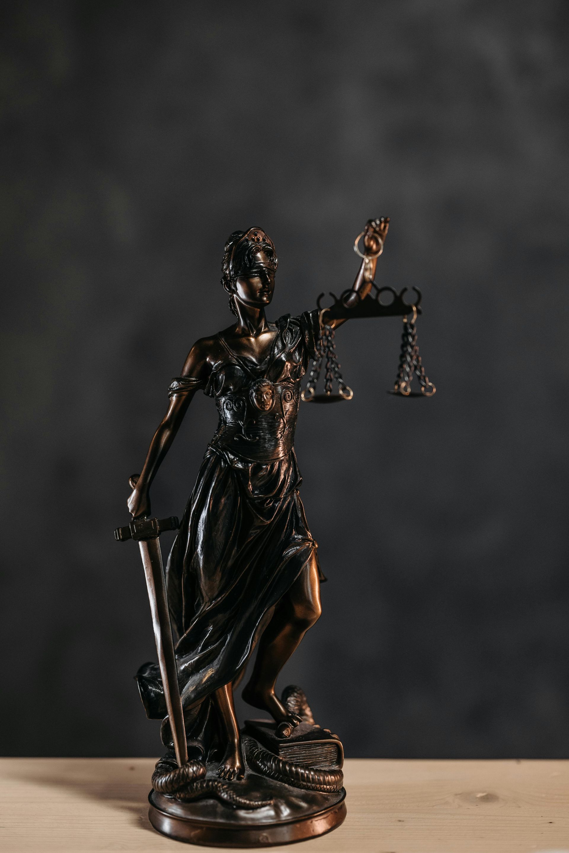 Statue of Lady Justice holding scales and a sword, bronze against a dark background.