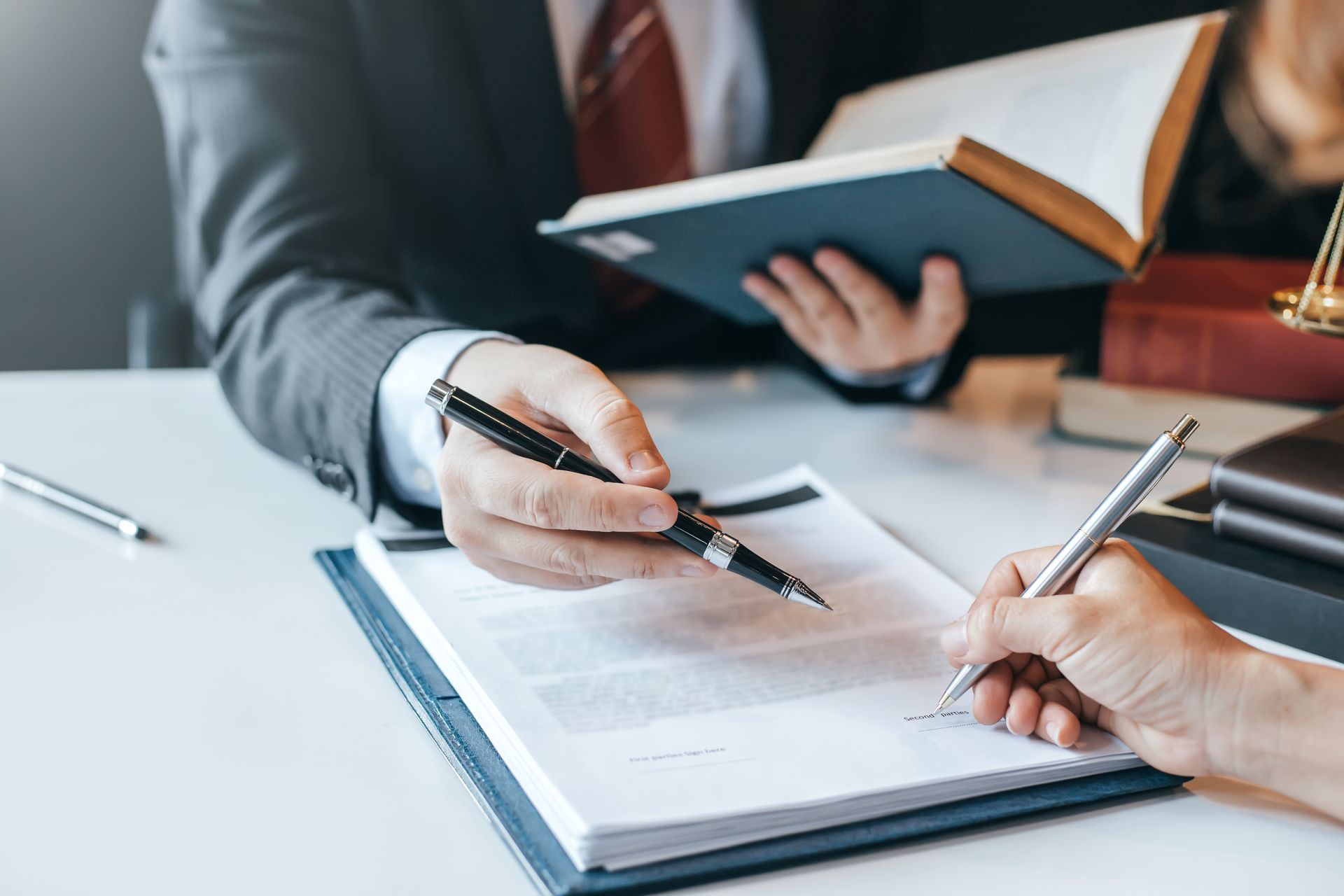 A lawyer points at a document as a person signs it, a book and scales of justice are nearby.