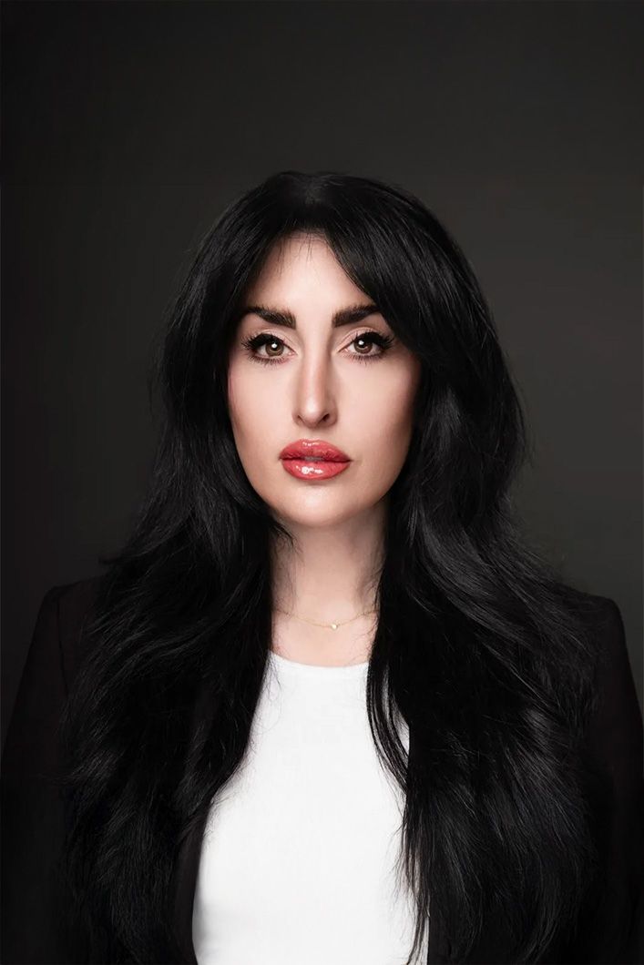 A woman with long black hair and red lips is wearing a white shirt and a black jacket.