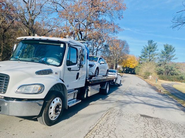 A white tow truck is carrying two cars down a road.