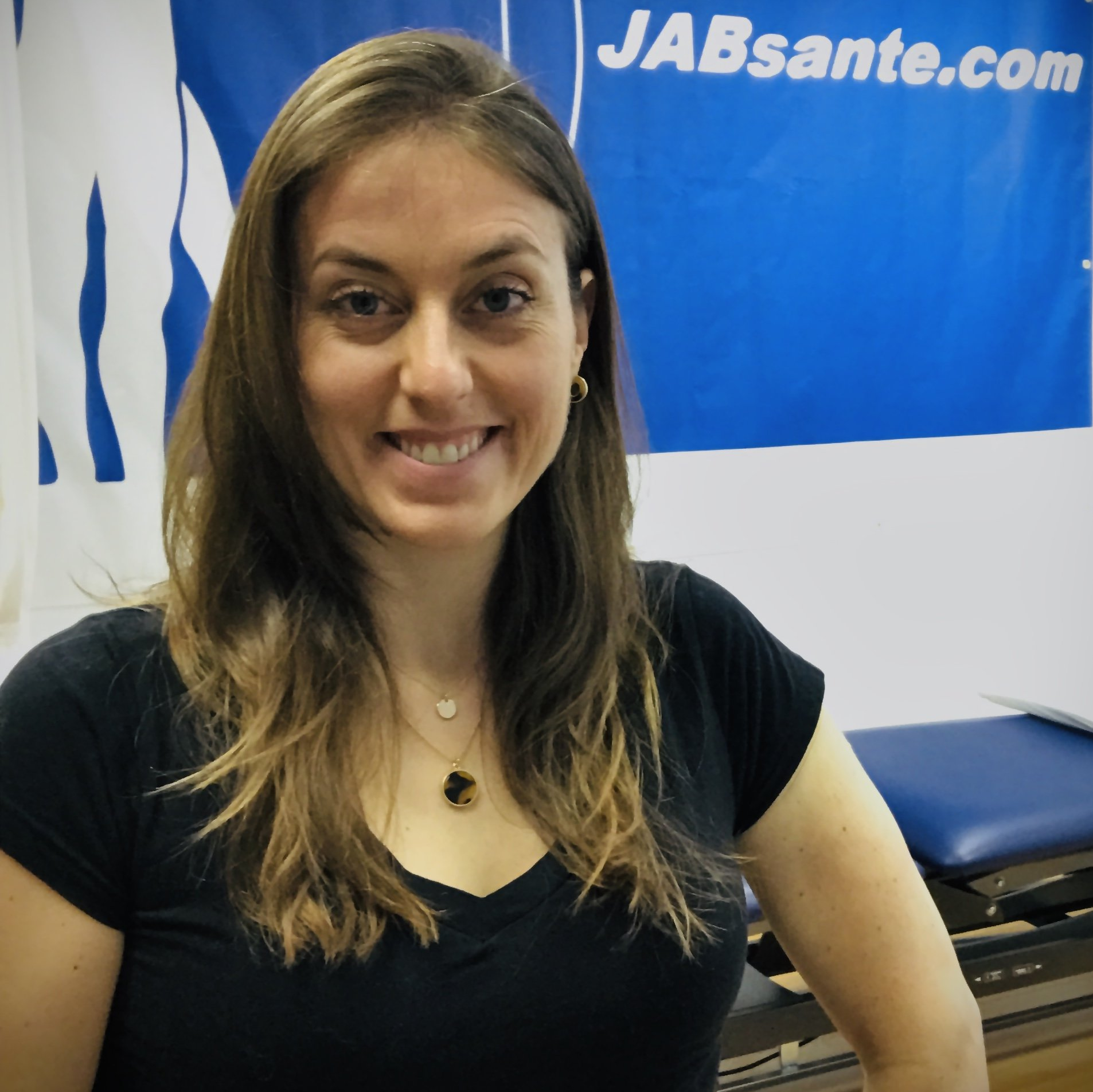 Woman smiling, standing in a clinic, wearing a black shirt, a blue backdrop with 