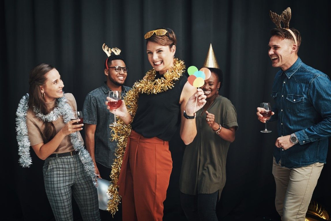 Group of friends celebrating, wearing party hats and tinsel, holding drinks and posing against a black background.