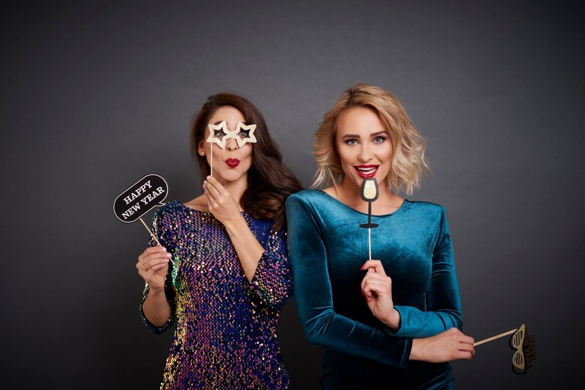 Two women pose with party props against a gray backdrop. One wears a sequined dress, star glasses, holding a