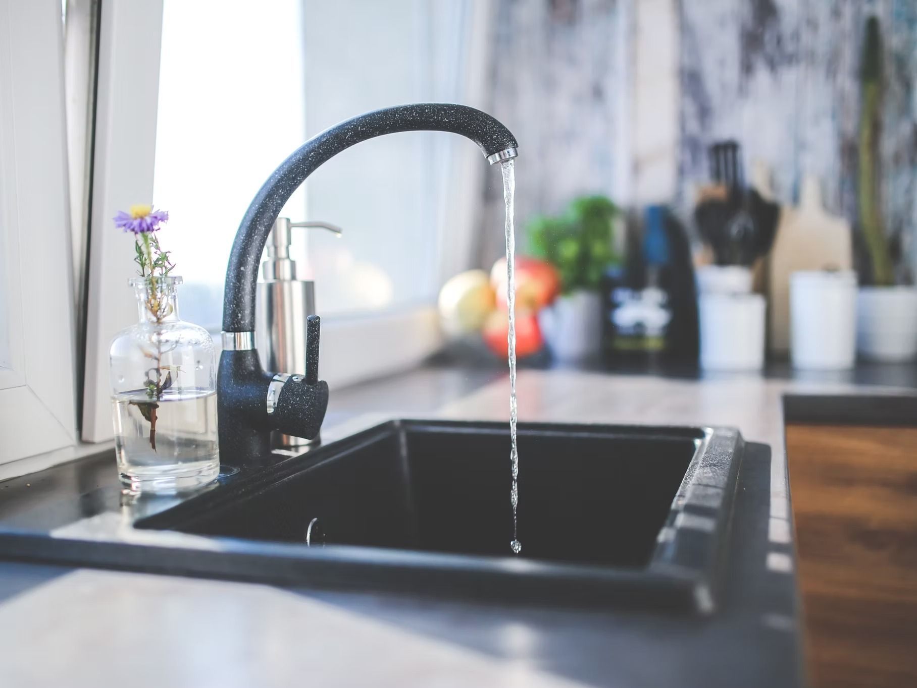Kitchen sink with running water; black sink, gray countertop, decorative items on windowsill and background.
