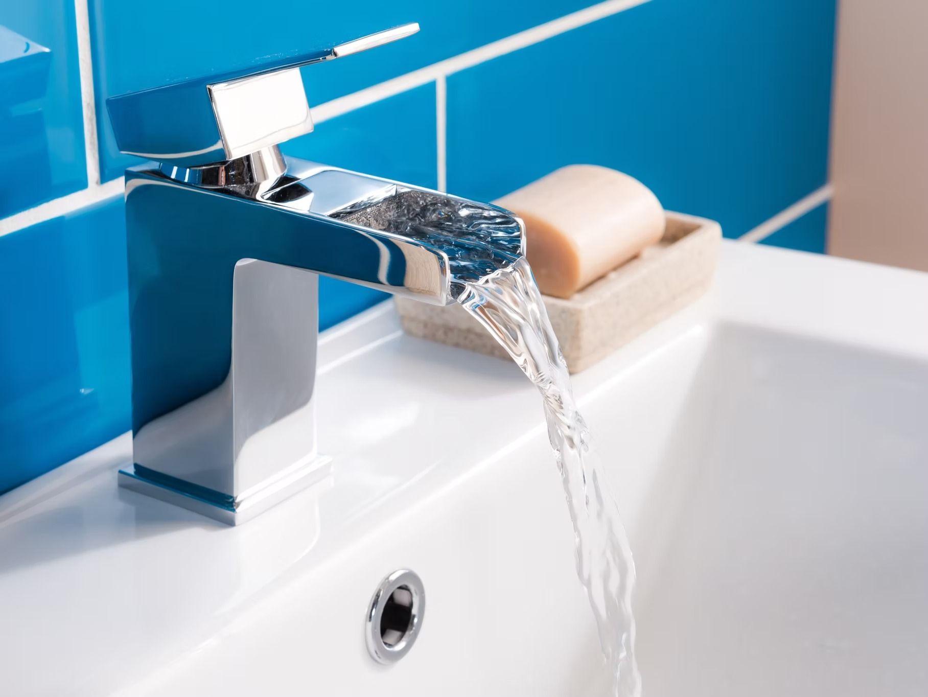 Chrome faucet pouring water into a white sink, blue tiled wall in the background. A bar of soap rests on a dish.