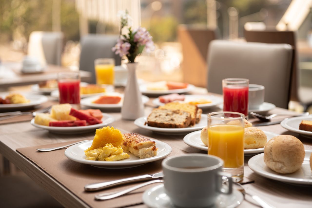 Buffet de café da manhã com frutas, sucos, ovos, pães e doces em uma mesa perto de uma janela.