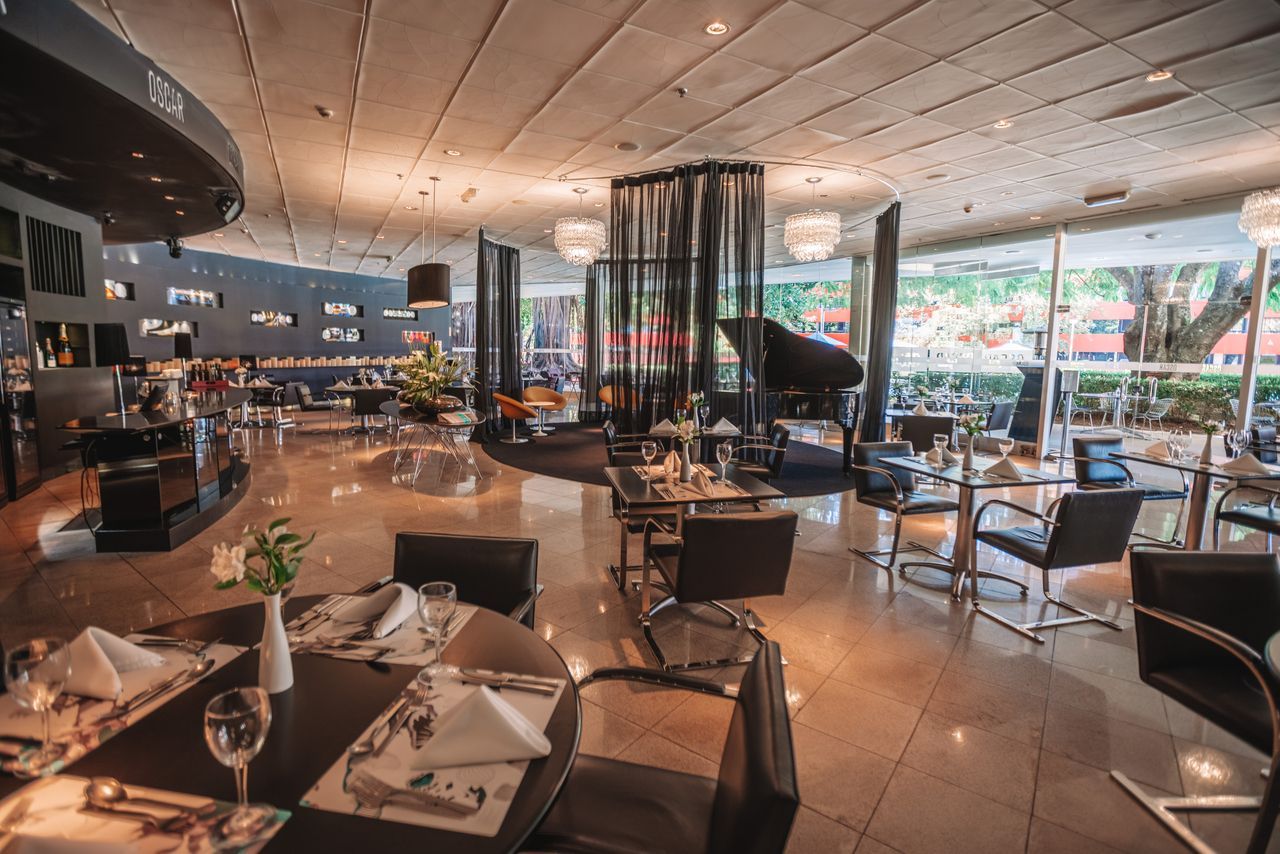 Interior elegante do restaurante com mesas, cadeiras e um piano de cauda perto de uma grande janela.