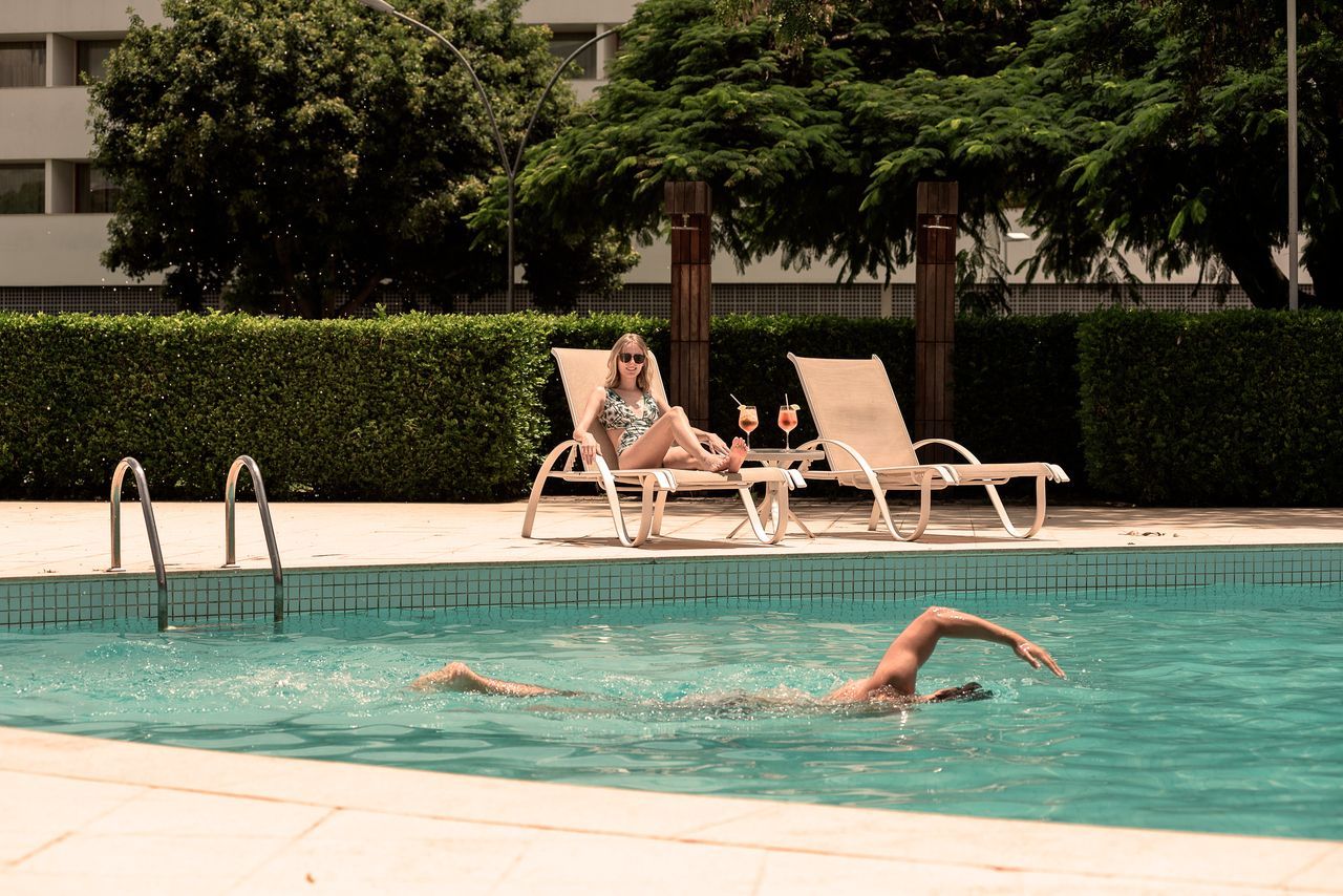Homem nadando livre na piscina, mulher relaxando na espreguiçadeira à beira da piscina, bebidas por perto.