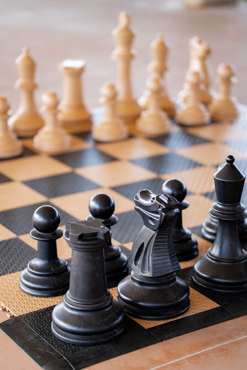Peças de xadrez em um tabuleiro. Peças pretas em foco: torre, cavalo, peão. Peças brancas fora de foco.