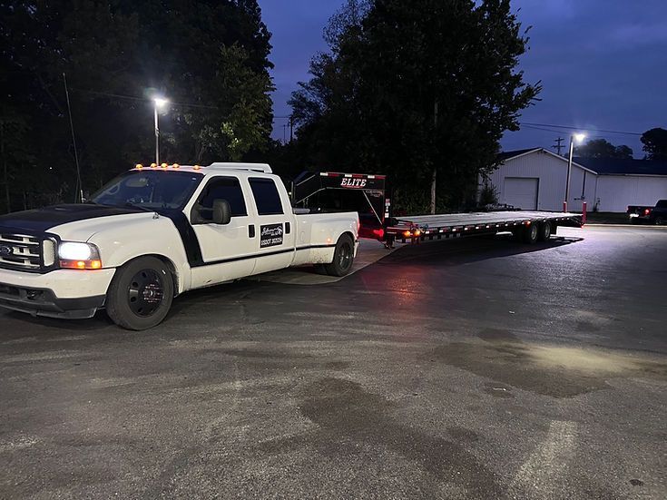 A white tow truck with a flatbed trailer attached to it is parked in a parking lot at night