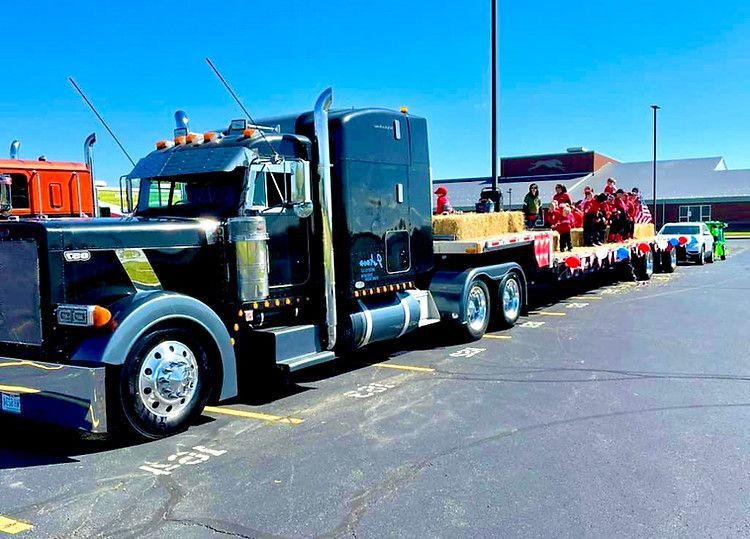 A large semi-truck is parked in a parking lot with a parade in the background