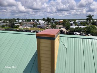 A chimney with a copper cap on a green metal roof, overlooking a neighborhood with palm trees under a cloudy sky.