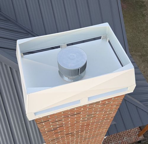 A white metal chimney shroud topped with a silver circular cap, mounted on a brick chimney against a dark metal roof.
