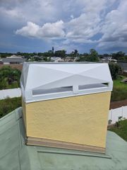 A textured yellow chimney with a white, angular metal cap sits on a green metal roof under a cloudy blue sky.
