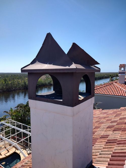 A metal chimney cap with a dual-gabled peak atop a white stucco chimney, set against a waterfront and tiled roof.