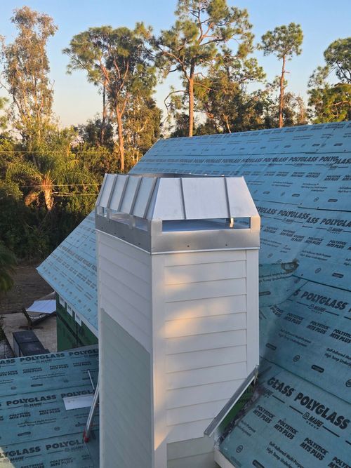 A white-sided chimney with a metal cap, situated on a sloped roof covered in blue underlayment against a treed backdrop.